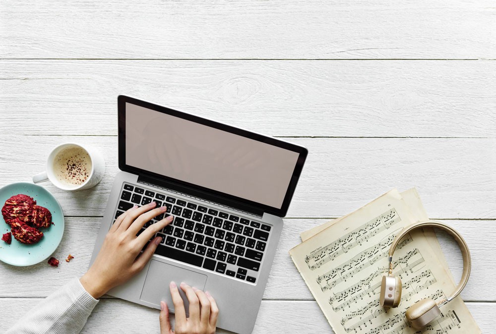 mesa de madera en la que una persona está escribiendo en un ordenador portátil Apple, partituras de música, un café y unas galletas de frambuesa